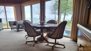 A dining area with a round table and four chairs, large windows with a view of the sea, and a wooden cabinet. - Lake Michigan Retreat (Menominee)