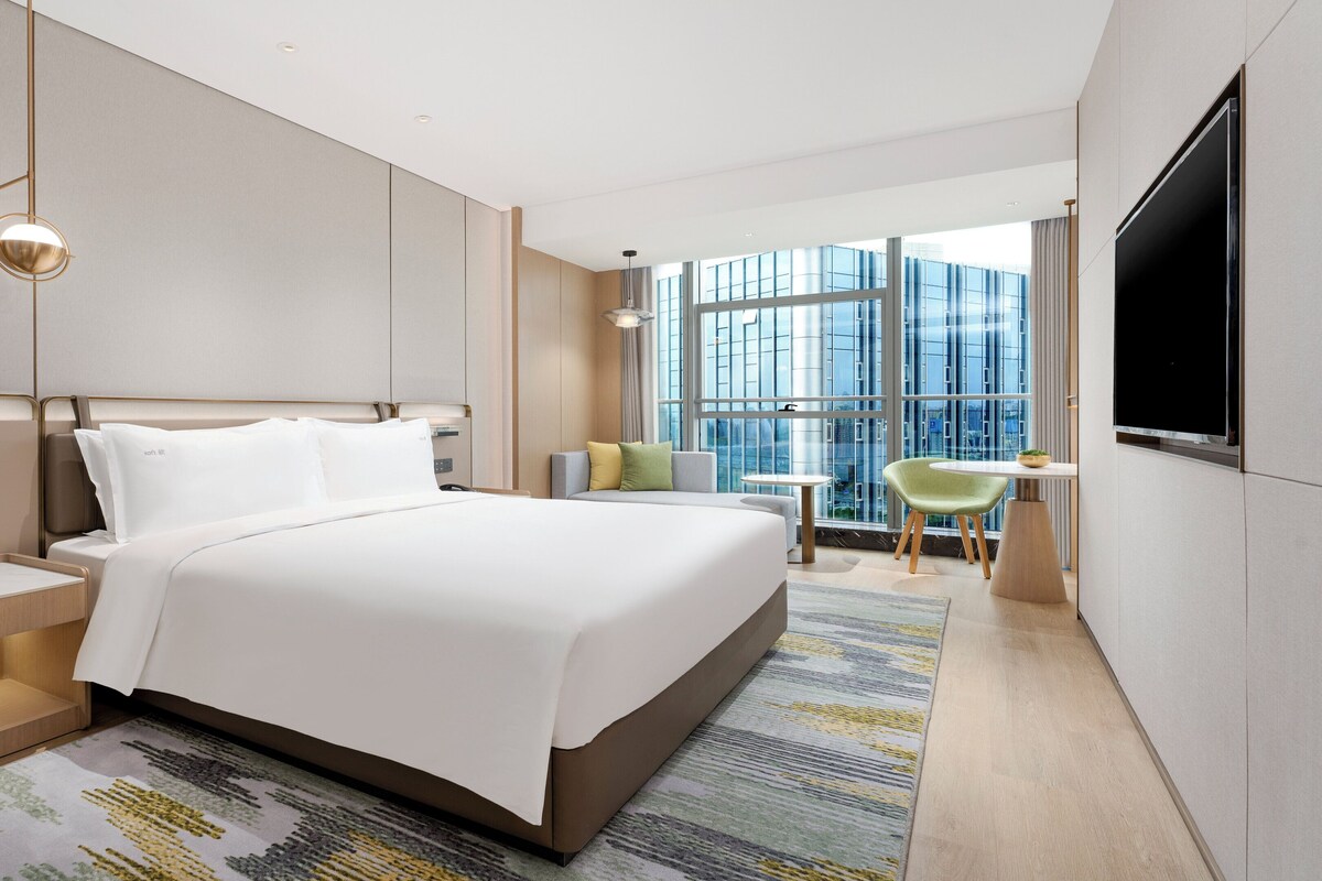A hotel room with a large bed, bedside table, a flat-screen TV, a seating area with a chair and table, and a large window offering a city view.