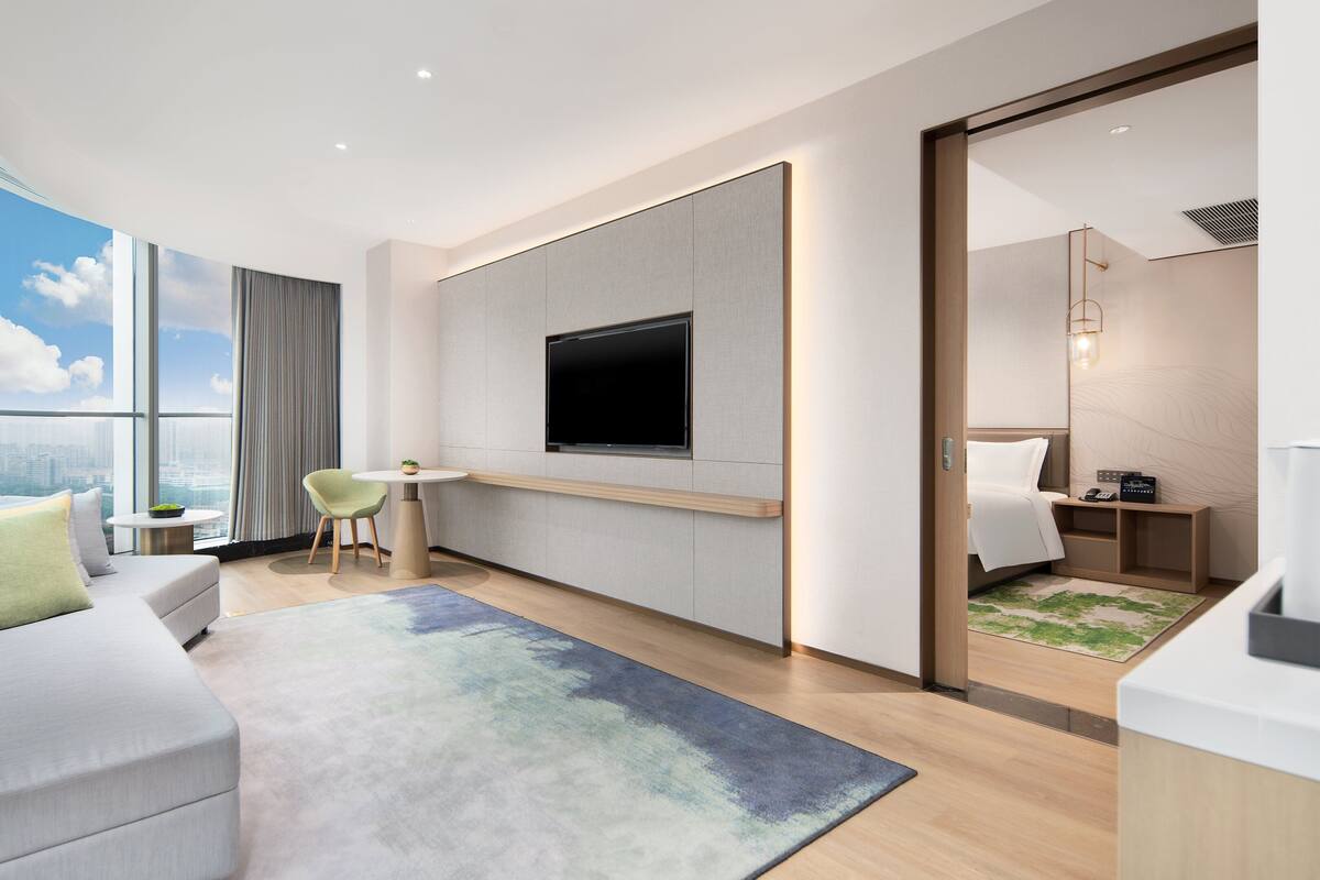 A modern hotel room with a large window, a flat-screen TV, a sofa, a small table, and a bed.