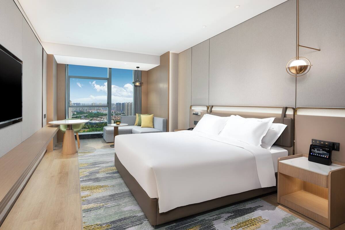 A modern hotel room with a large bed, a flat-screen TV, a desk, and a view of the city.
