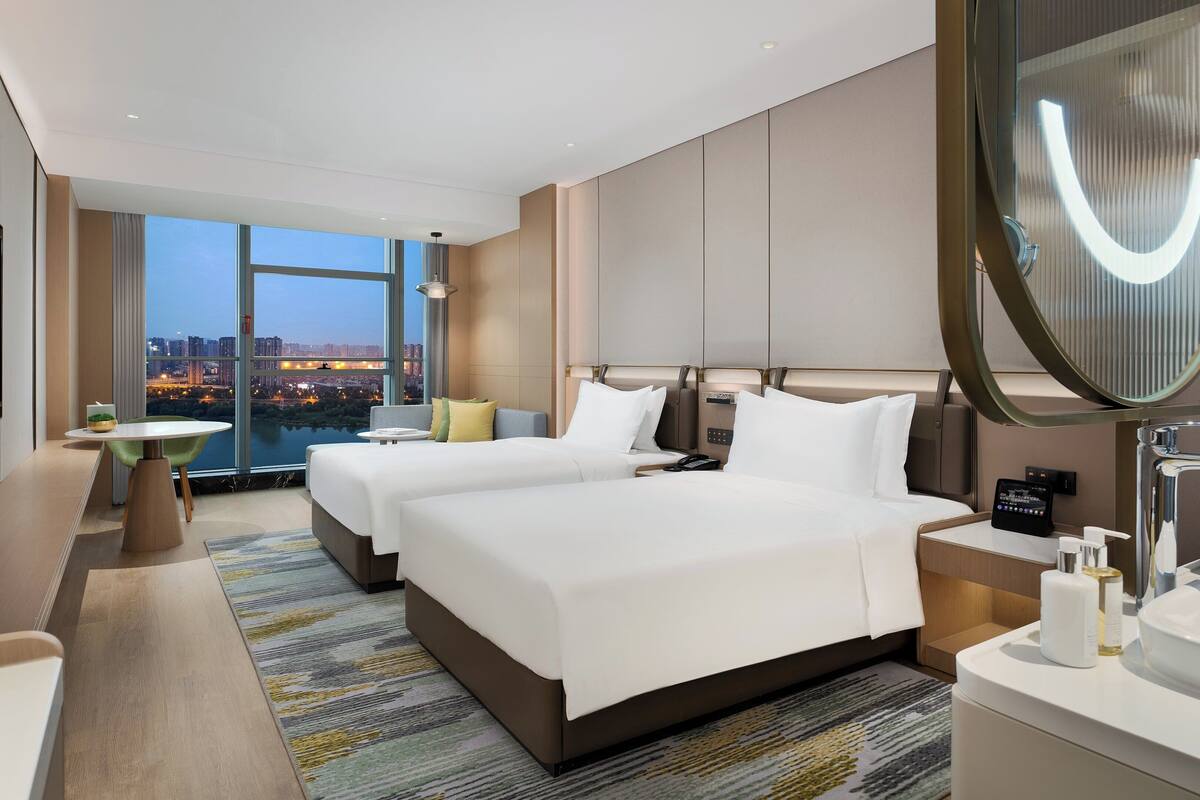 A modern hotel room with a large bed, a desk, a chair, a small table, a TV, a bathroom with a mirror, and a view of the cityscape.