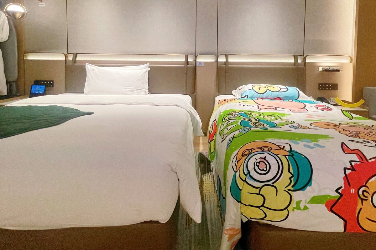 Een hotelkamer met twee bedden, waarvan één een effen witte spreidek en de ander een kleurrijke bedspreide met tekenfilms.
