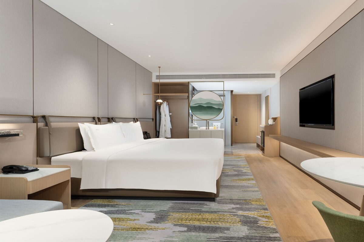 A modern hotel room with a large bed, a desk with a telephone, a round table with a bowl of greenery, and a television mounted on the wall.