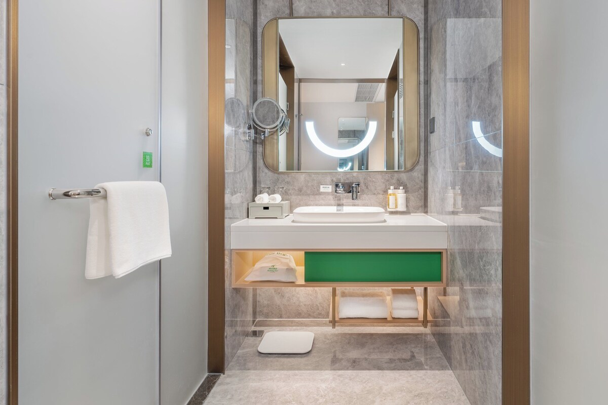 Een moderne badkamer met een douchecabine van glas, een witte wastafel met een groene kast, en een grote spiegel met een ronde lamp erboven.