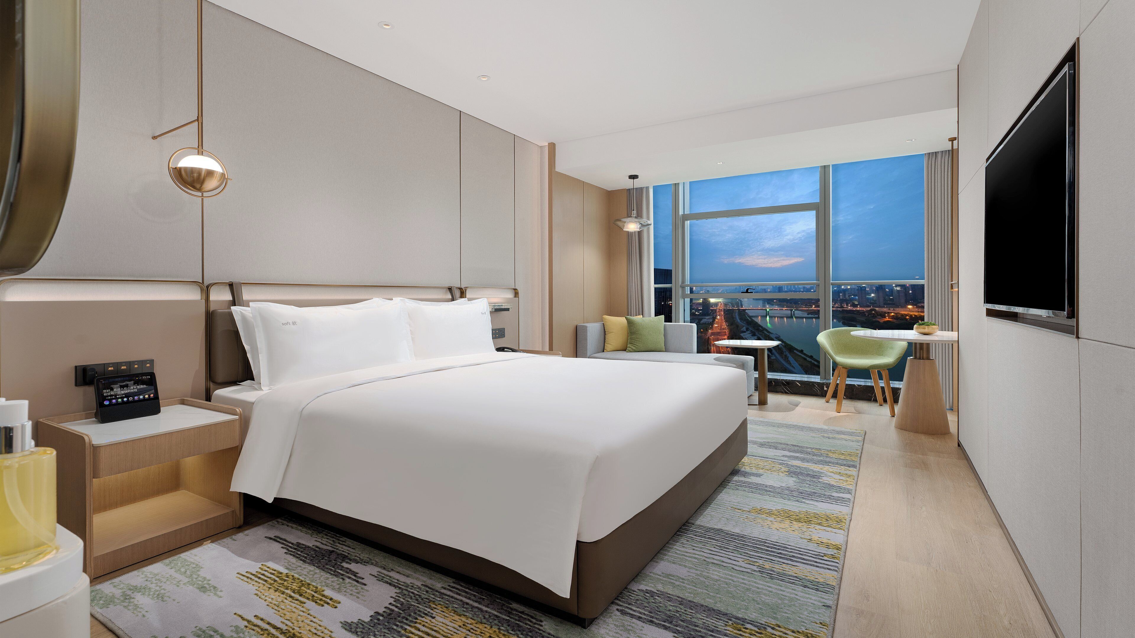 A modern hotel room with a large bed, a desk, a TV, and a view of a cityscape.