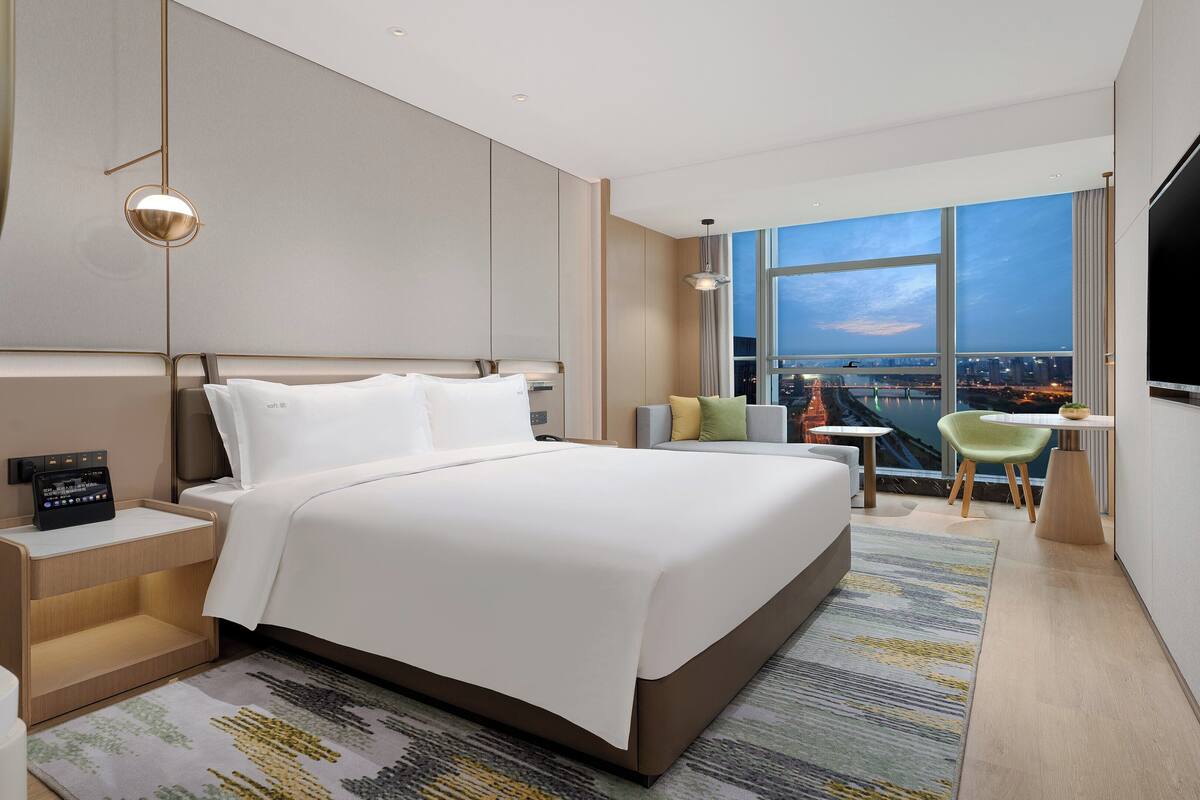 A modern hotel room with a large bed, a desk, a TV, and a view of a cityscape.