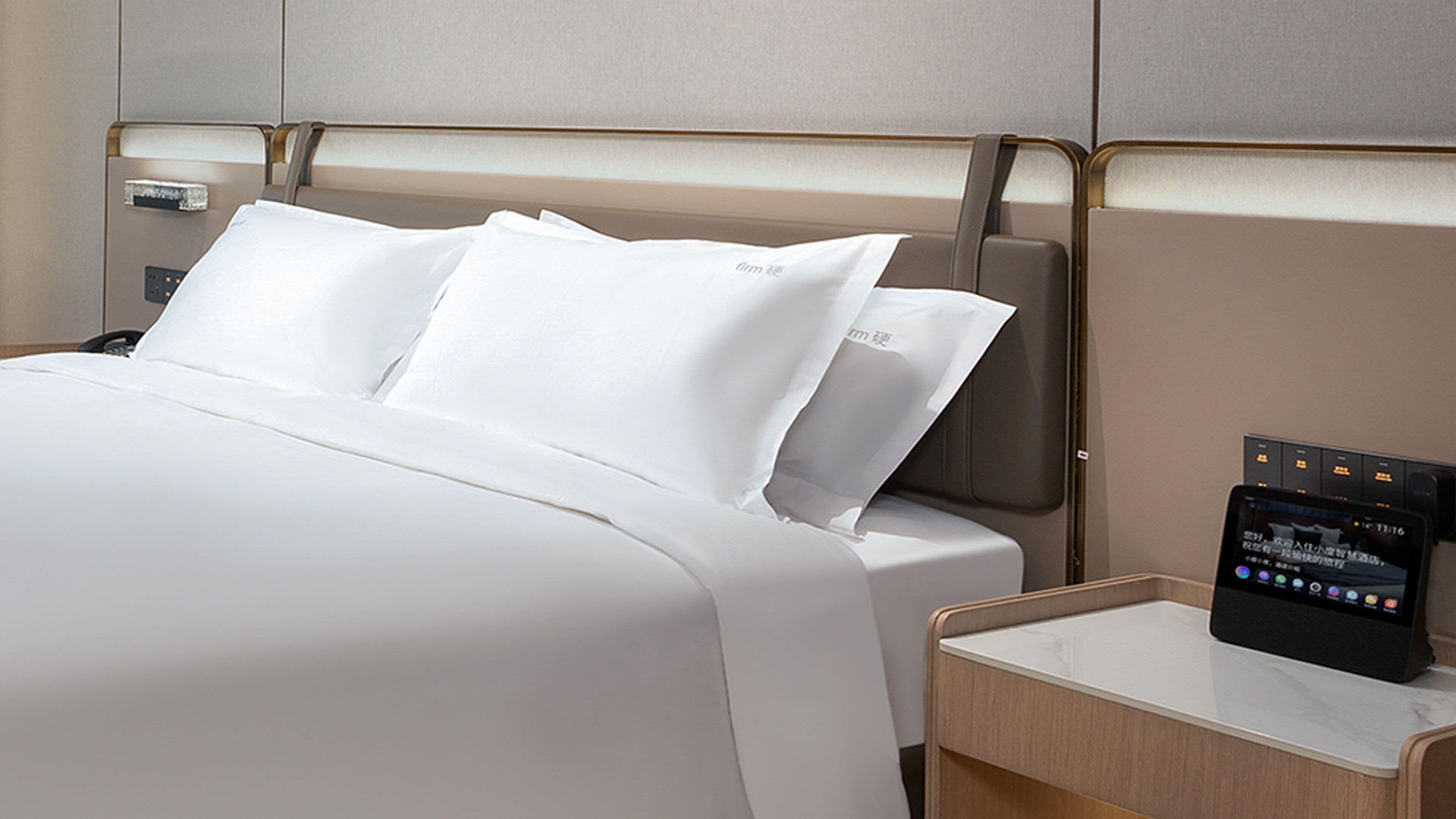 A hotel room with a neatly made bed, a bedside table with a digital control panel, and a wall-mounted panel.