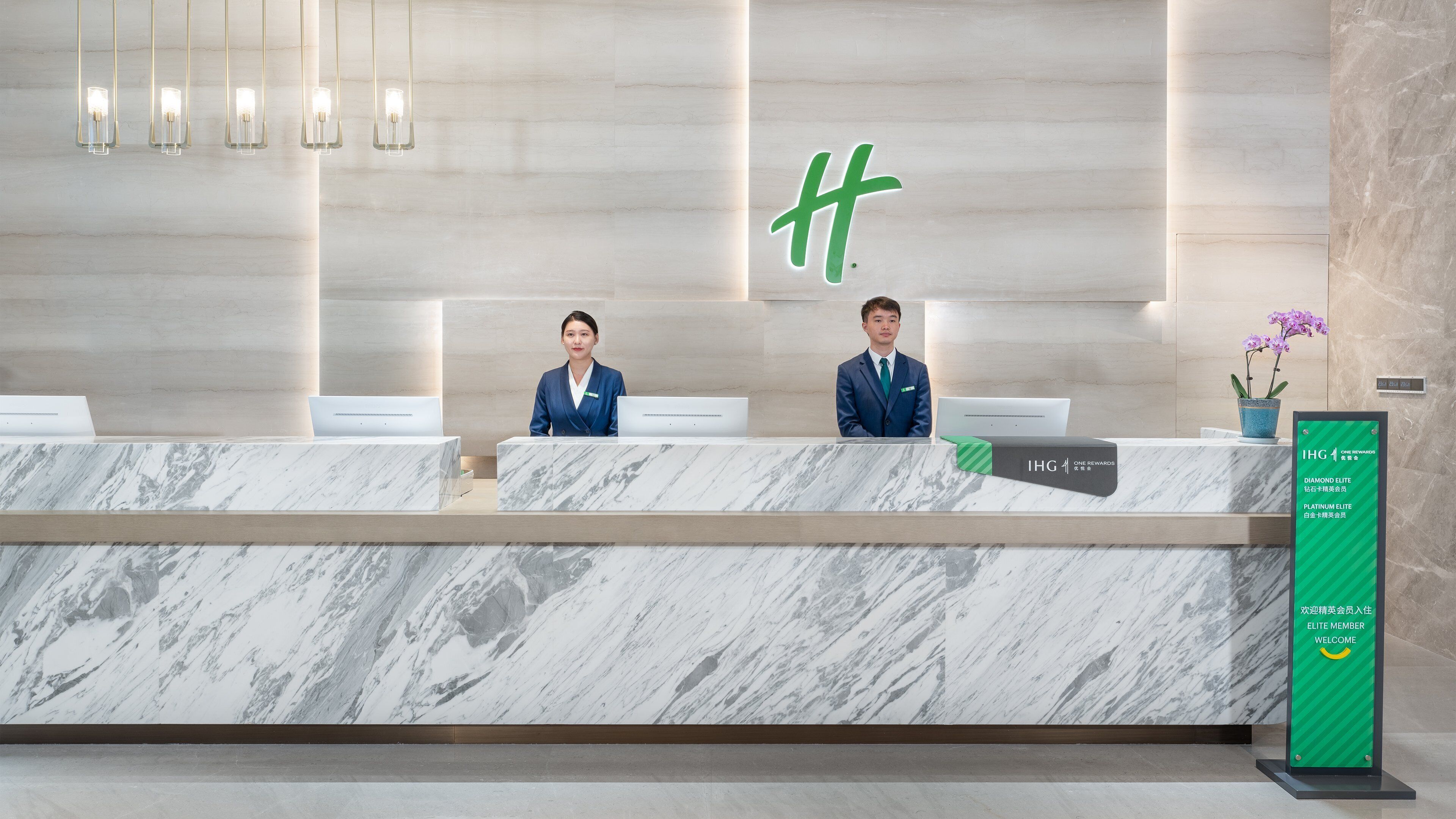 En hotelreception med marmorbordplader, to receptionister og et skilt, der viser hotellets faciliteter.
