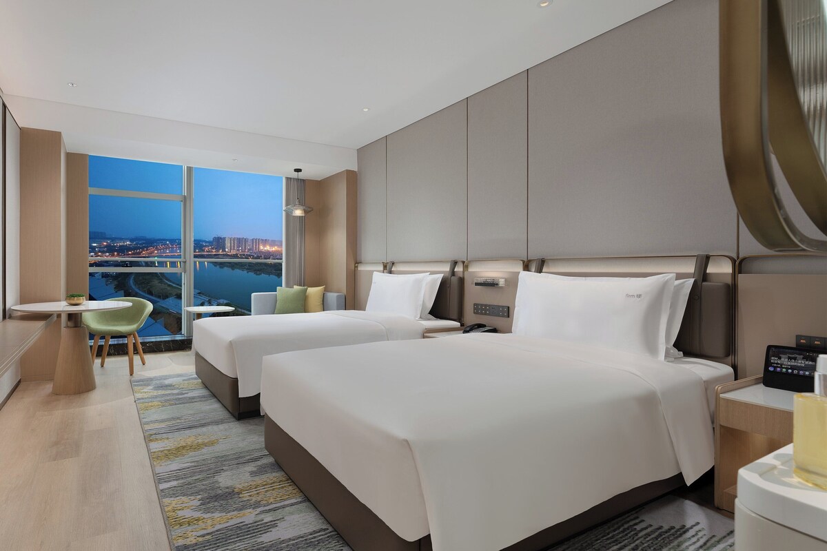 A hotel room with two beds, a TV, a small table, and a view of a cityscape.