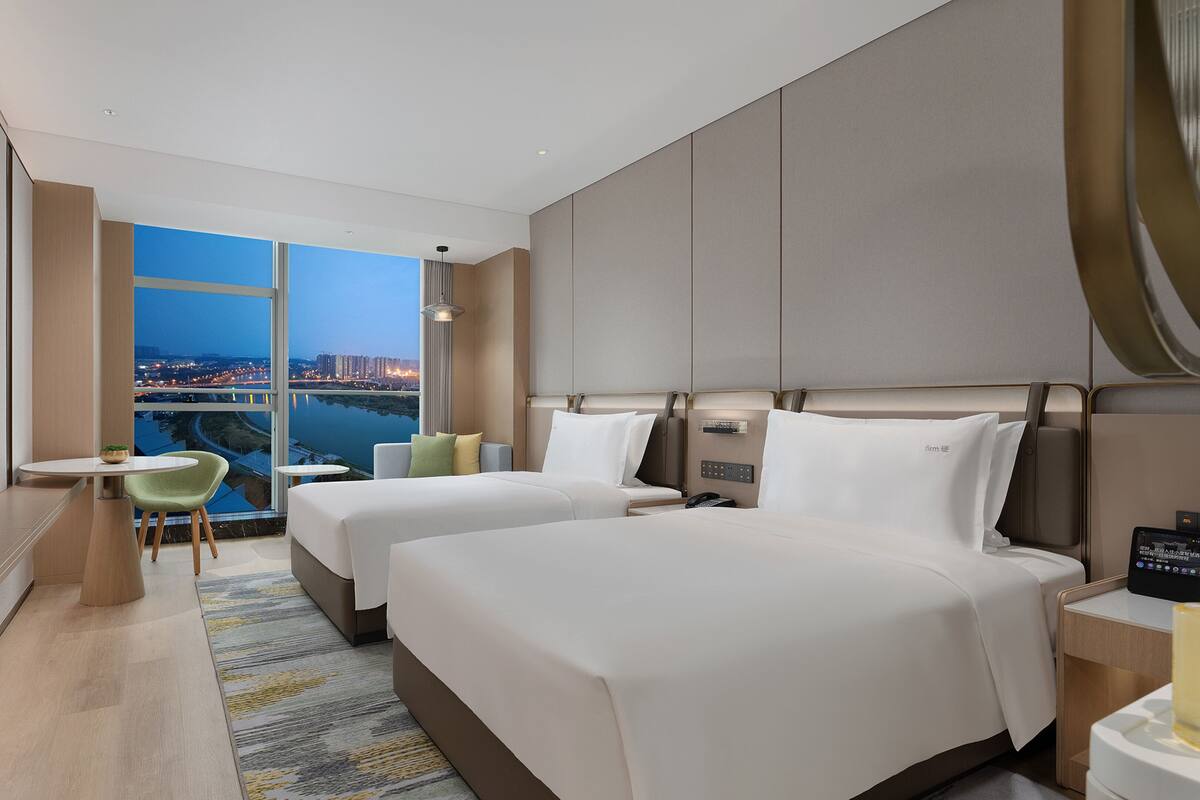 A hotel room with two beds, a TV, a desk, and a view of a cityscape.