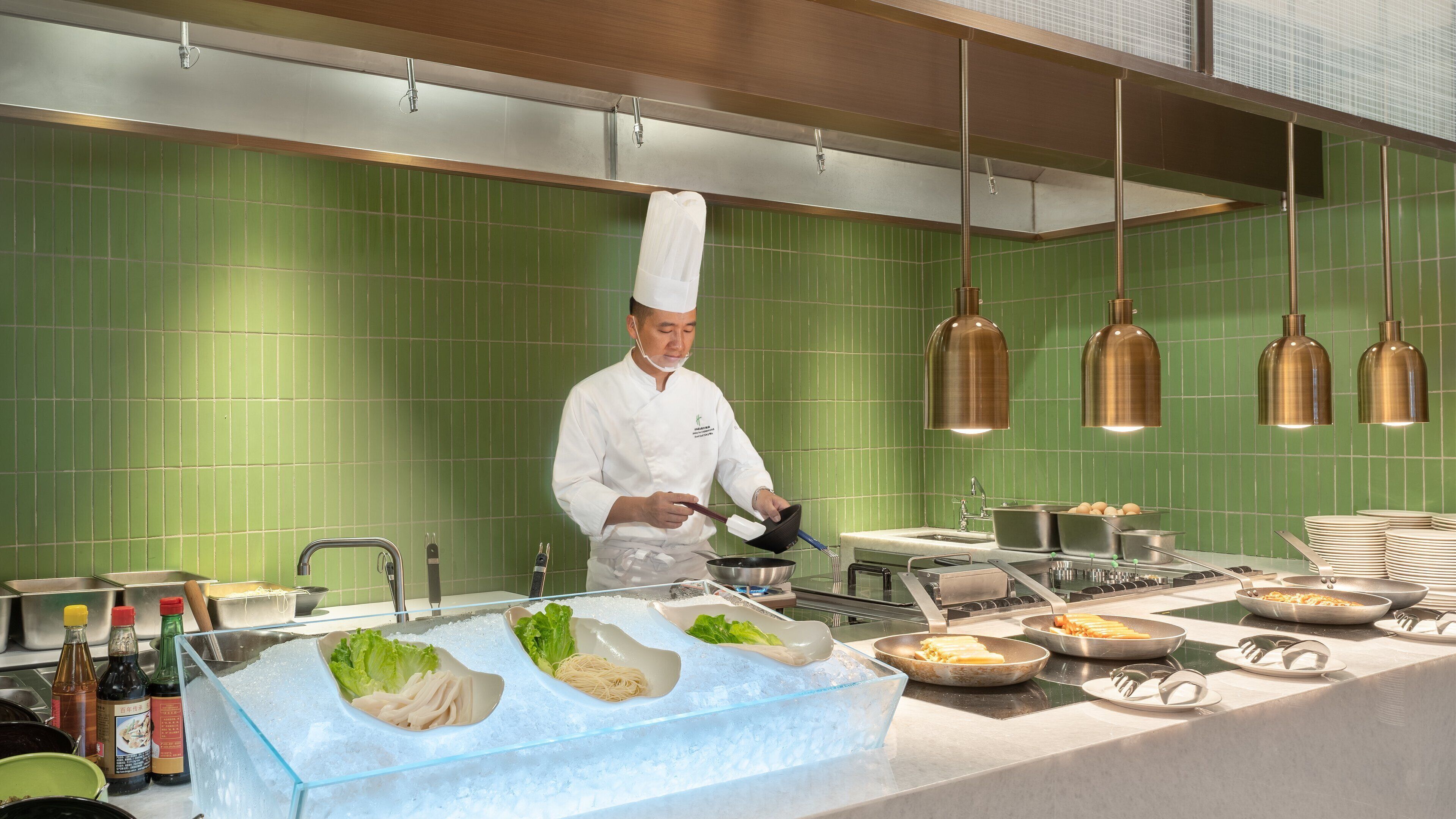 A chef in a kitchen with green tiled walls, stainless steel appliances, and a variety of food items on display.