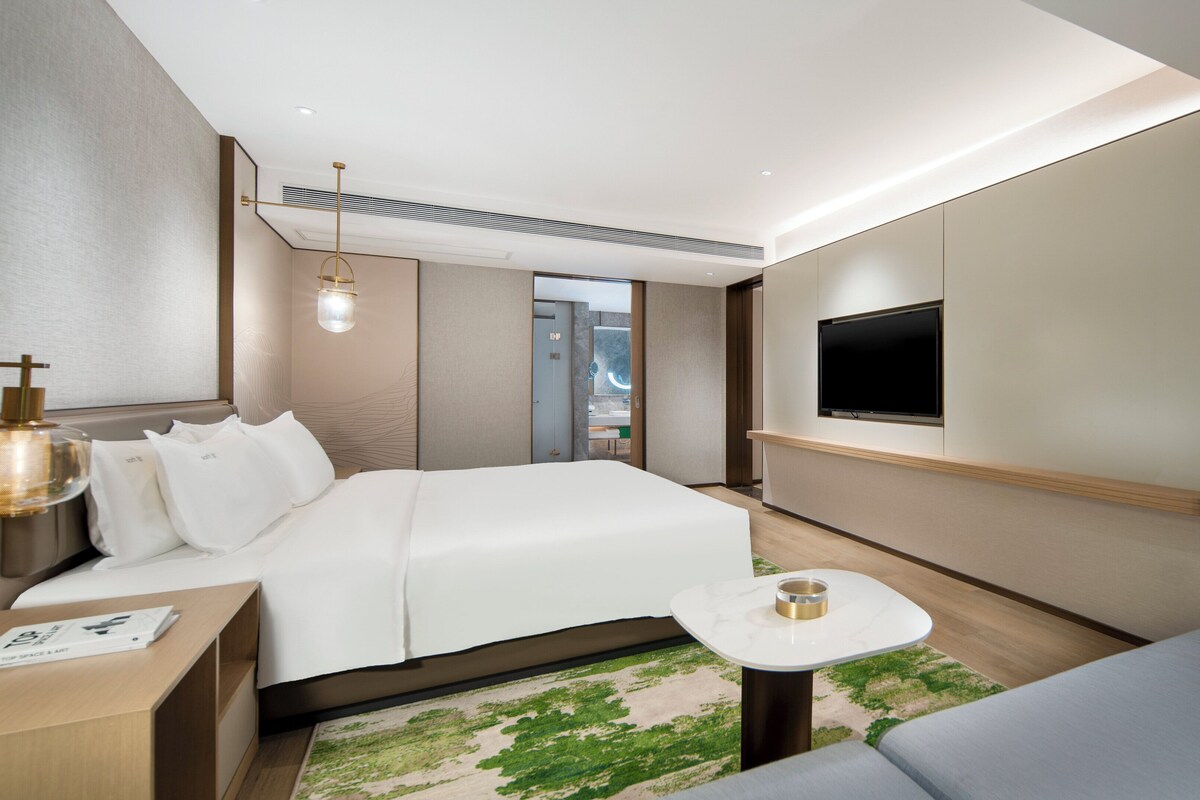 Een moderne hotelkamer met een netjes opgemaakt bed, een nachtkastje met een lamp, een flatscreen televisie aan de muur en een klein rond tafeltje met een kop.