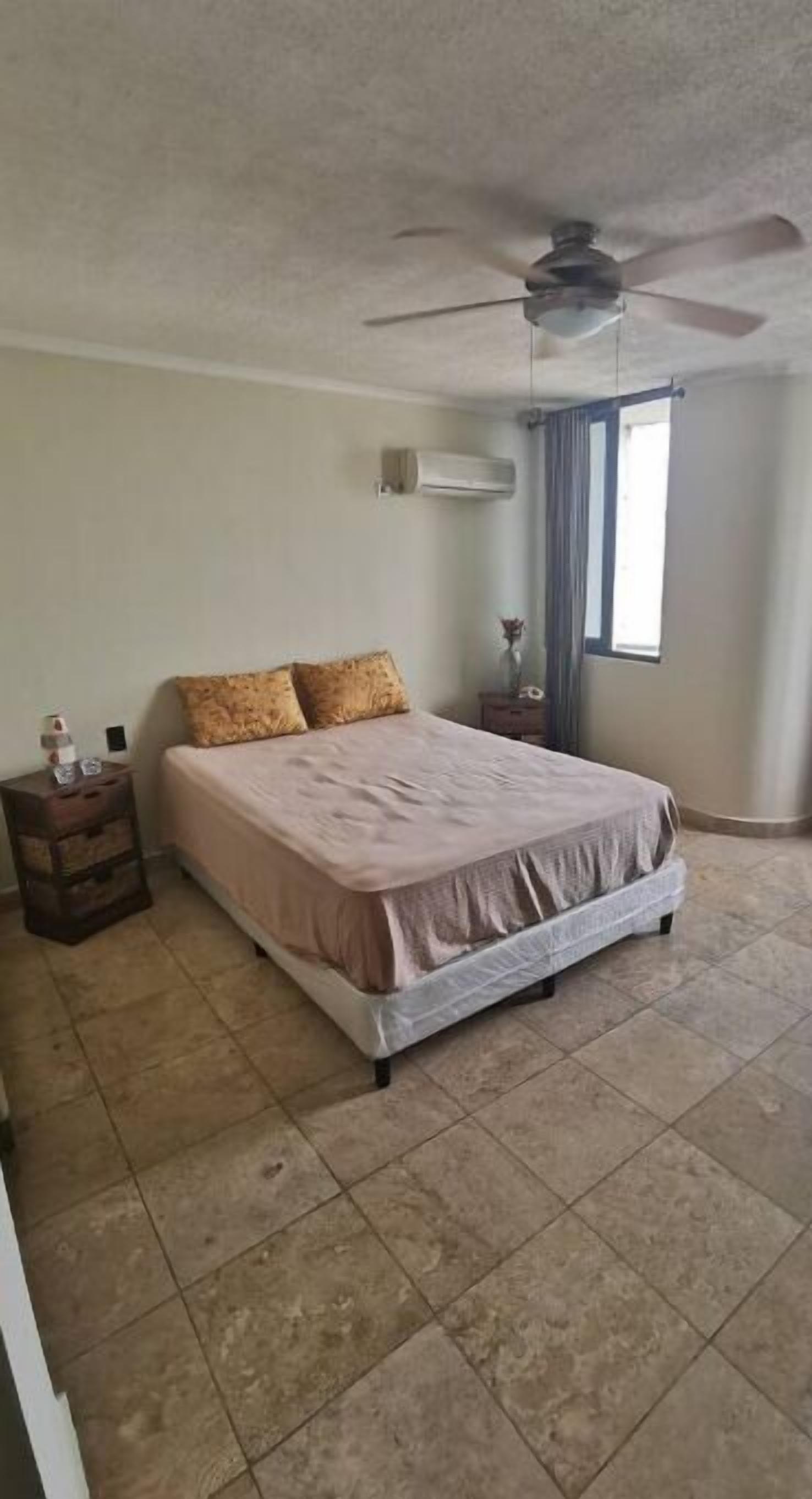 A bedroom with a bed, a ceiling fan, an air conditioning unit, a nightstand, a window with curtains, and a tiled floor.