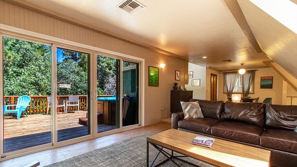 A spacious living room with a brown leather sofa, a wooden coffee table, and a sliding glass door leading to an outdoor deck with a blue chair and a wooden table. - Hot Tub Hideaway  (Idyllwild-Pine Cove)