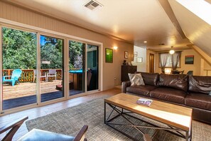 Un salon spacieux avec un canapé en cuir marron, une table basse en bois et une porte-fenêtre coulissante donnant sur une terrasse extérieure équipée d’une chaise bleue et d’une table en bois.