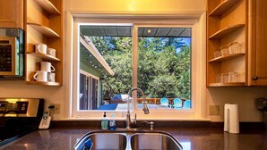 A kitchen with a double sink, wooden cabinets, and a window overlooking a deck. - Hot Tub Hideaway  (Idyllwild-Pine Cove)