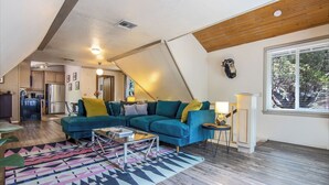 A modern living room with a blue sectional sofa, a glass coffee table, and a colorful rug. A kitchen is visible in the background, and there is a large window providing natural light. - Hot Tub Hideaway  (Idyllwild-Pine Cove)
