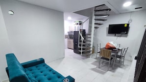 Una sala de estar con un sofá color turquesa, una mesa de comedor con sillas, un televisor montado en la pared y una escalera que sube a un nivel superior.