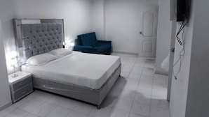 Un dormitorio con una cama grande, una mesita de noche, una silla azul y un televisor fijado a la pared.