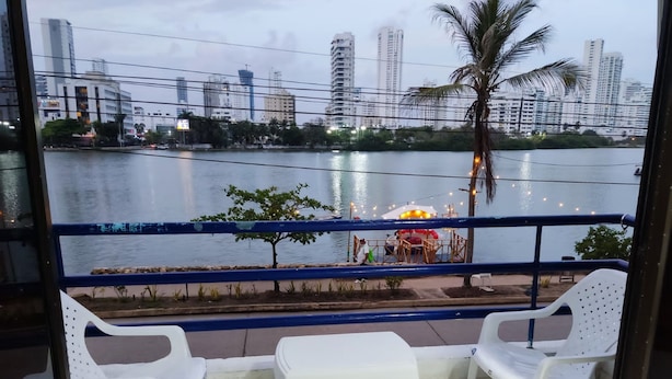 Un balcón con sillas de plástico blanco, baranda y vista a un río con edificios y una palmera al fondo.