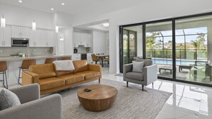 A modern kitchen with white cabinets, a dining area with a table and chairs, a living room with a sofa and armchairs, and a large sliding glass door leading to a pool area. - Brand NEW Construction, Beautiful Home, HEATED Pool in Highly Desirable SW Cape! (Cape Coral)