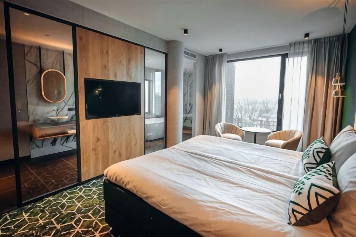 A modern hotel room with a large bed, a TV, a bathroom with a sink and mirror, and a view of the city.