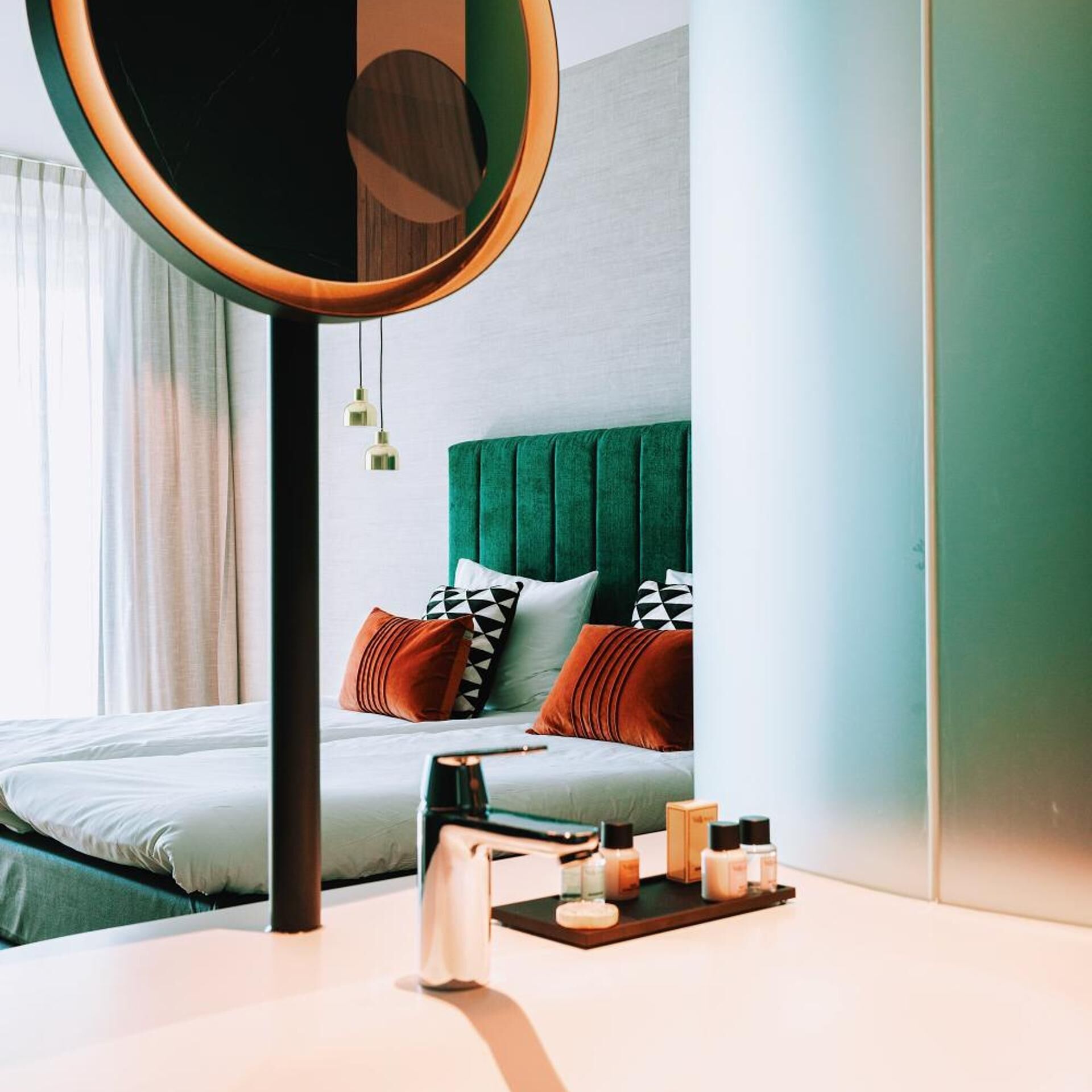 Une chambre d’hôtel moderne avec un lit, une table de chevet avec des articles de toilette, un miroir rond et une tête de lit recouverte d’un tissu vert.