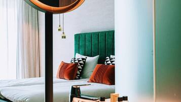 Une chambre d’hôtel moderne avec un lit, une table de chevet avec des articles de toilette, un miroir rond et une tête de lit recouverte d’un tissu vert.