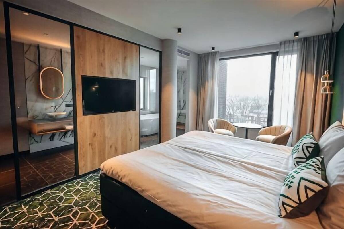 A modern hotel room with a large bed, a TV, a bathroom with a sink and mirror, and a view of the city.