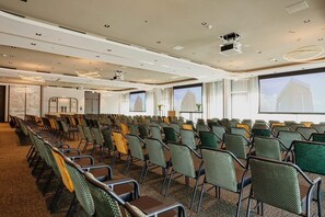 A conference room with rows of chairs, a projector screen, and a large window with a view of a building. - Van der Valk Hotel Schiedam (Schiedam)