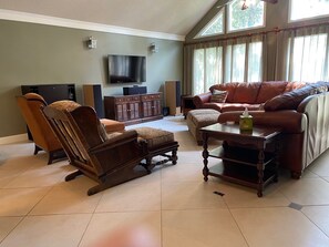 Un salon avec un canapé en cuir marron, un fauteuil à bascule en bois et un pouf assorti. Un téléviseur à écran plat est fixé au mur, et deux haut-parleurs sont placés de chaque côté.