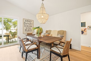 Een eetgedeelte met een houten tafel, rieten stoelen, een kroonluchter en een groot raam met uitzicht op de omgeving.