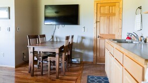 Een eetgedeelte met een houten tafel en stoelen, een aan de muur bevestigde televisie, een houten deur en een keuken met een gootsteen en aanrecht.