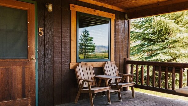 Een houten veranda met twee Adirondack-stoelen en een tafel, een deur met het nummer 5, en een raam met uitzicht op bomen en bergen.
