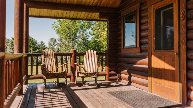 A wooden cabin with a porch featuring two Adirondack chairs and a wooden door with a window.