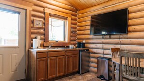 A log cabin interior with a kitchen area featuring wooden cabinets, a sink, a dishwasher, a coffee maker, and a television mounted on the wall.