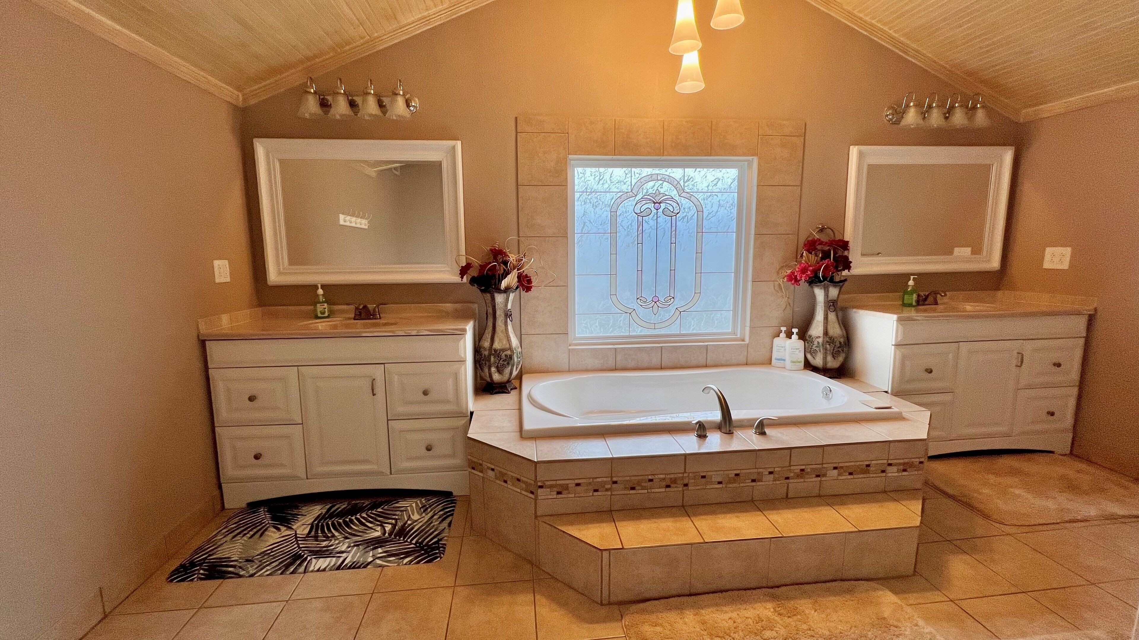 Une salle de bain spacieuse avec une grande baignoire, deux lavabos et un meuble-lavabo avec des placards. On y trouve également des vases décoratifs et un tapis sur le sol.