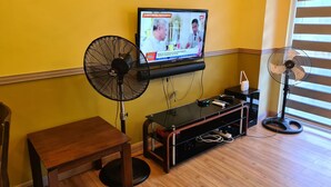 A room with a television mounted on the wall, two standing fans, a wooden table, and a black entertainment unit. - PICO DE LORO ROOM FOR RENT by BILLYN HAMILO COAST, BATANGAS (Nasugbu)