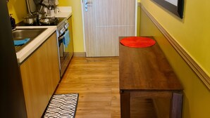 A compact kitchen with wooden cabinets, a microwave, and a stove. There is a wooden table with a red tray and a trash bin. - PICO DE LORO ROOM FOR RENT by BILLYN HAMILO COAST, BATANGAS (Nasugbu)