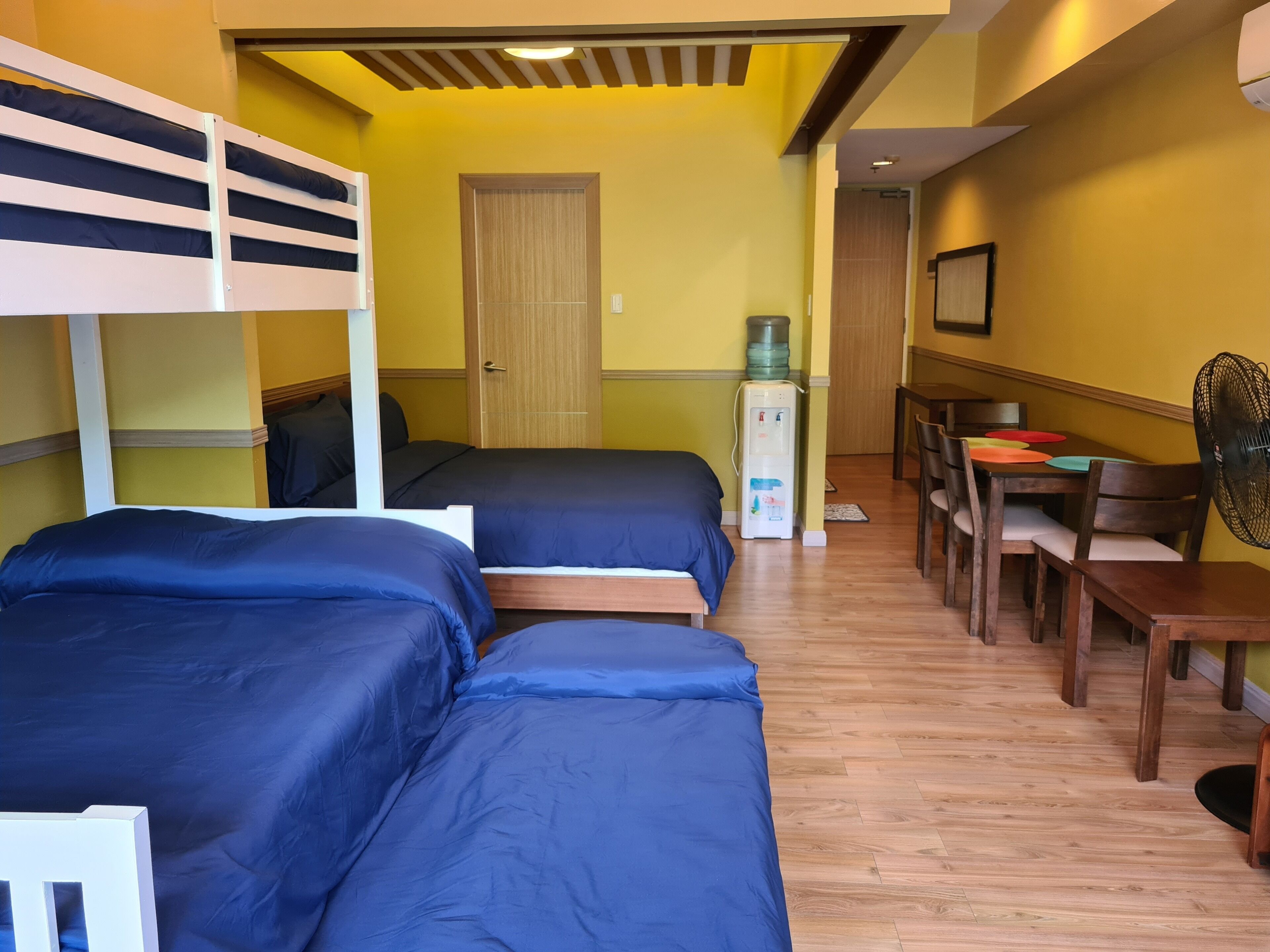 Een hotelkamer met stapelbedden, een eettafel met stoelen, een waterdispenser en een ventilator.