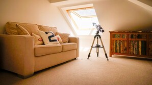 Une pièce mansardée avec un plafond en pente, un télescope sur un trépied, un canapé beige avec des coussins et un meuble en bois.
