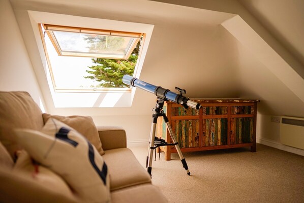 A telescope on a tripod in a room with a sloped ceiling and a large window.