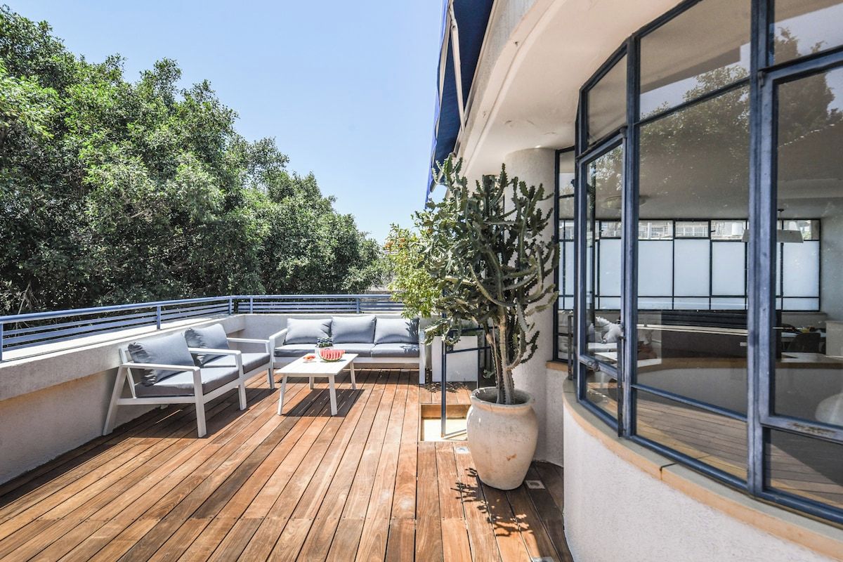 Une terrasse extérieure avec un plancher en bois, des meubles blancs et une grande plante en pot.