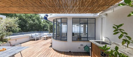 A covered outdoor deck with a kitchen area, seating, and a view of the surrounding greenery.