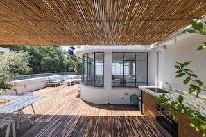 A covered outdoor deck with a kitchen area, seating, and a view of the surrounding greenery.