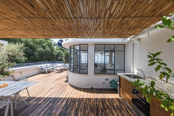 Une terrasse extérieure couverte, dotée d’un espace cuisine, de sièges et offrant une vue sur la verdure environnante.