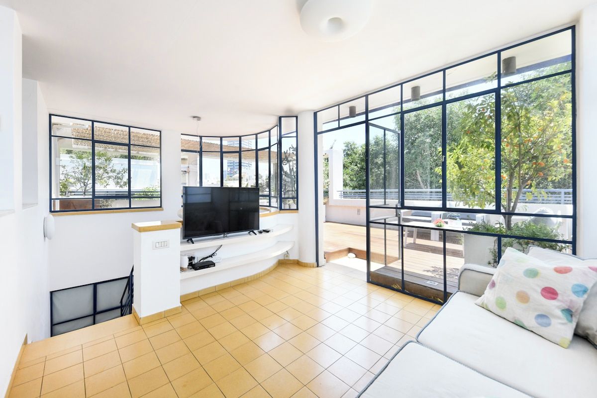 Ein modernes Wohnzimmer mit Fernseher, großen Fenstern und einem weißen Sofa.