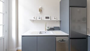 A modern kitchen with a stainless steel refrigerator, a double sink, and a dishwasher. There are wall-mounted shelves with framed pictures and a light fixture above.