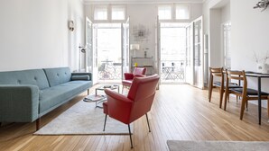 A spacious living area with a sofa, chairs, a dining table, and a view through open doors to the outside.