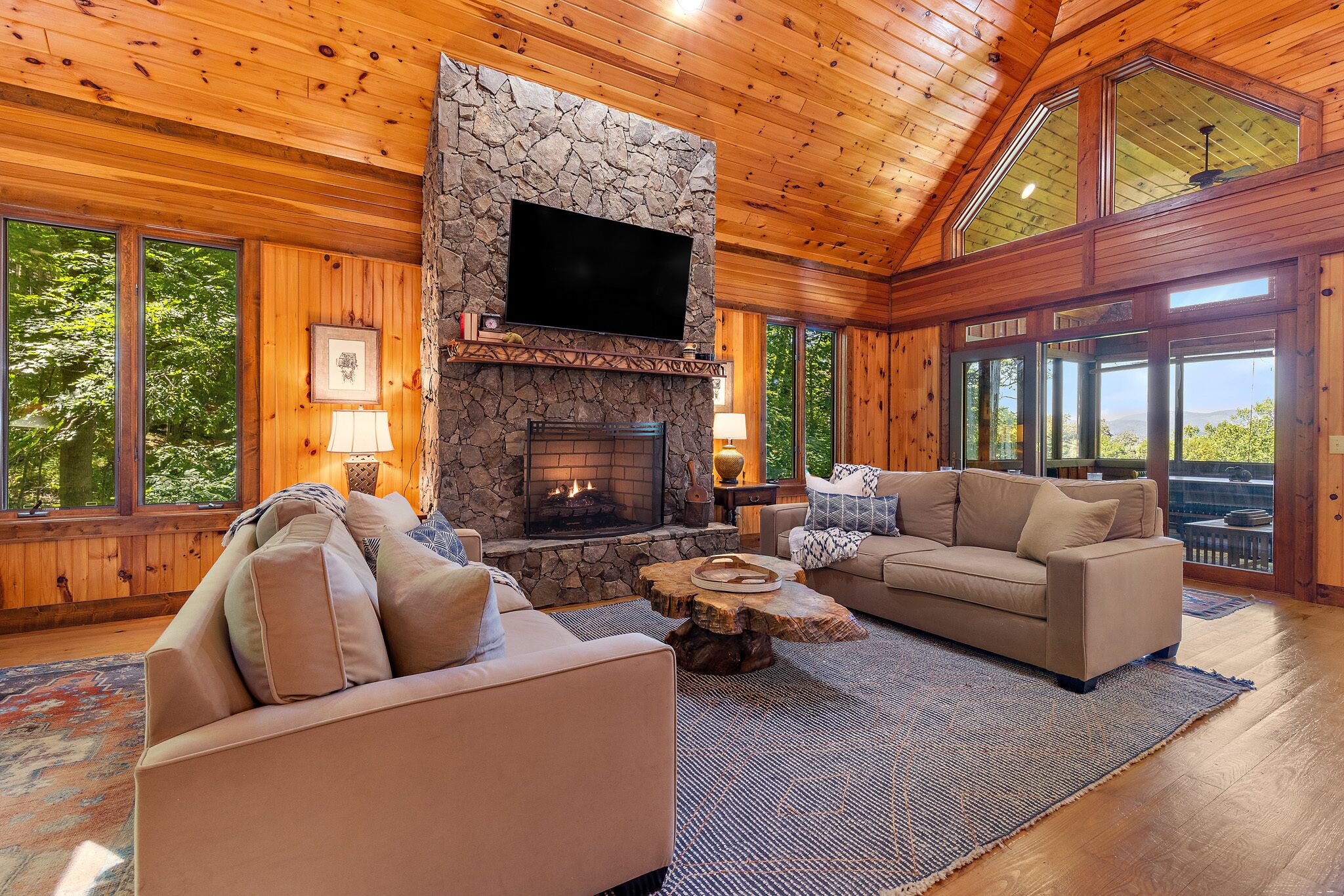 A cozy living room with a stone fireplace, wooden ceiling, and large windows offering a view of the outdoors.